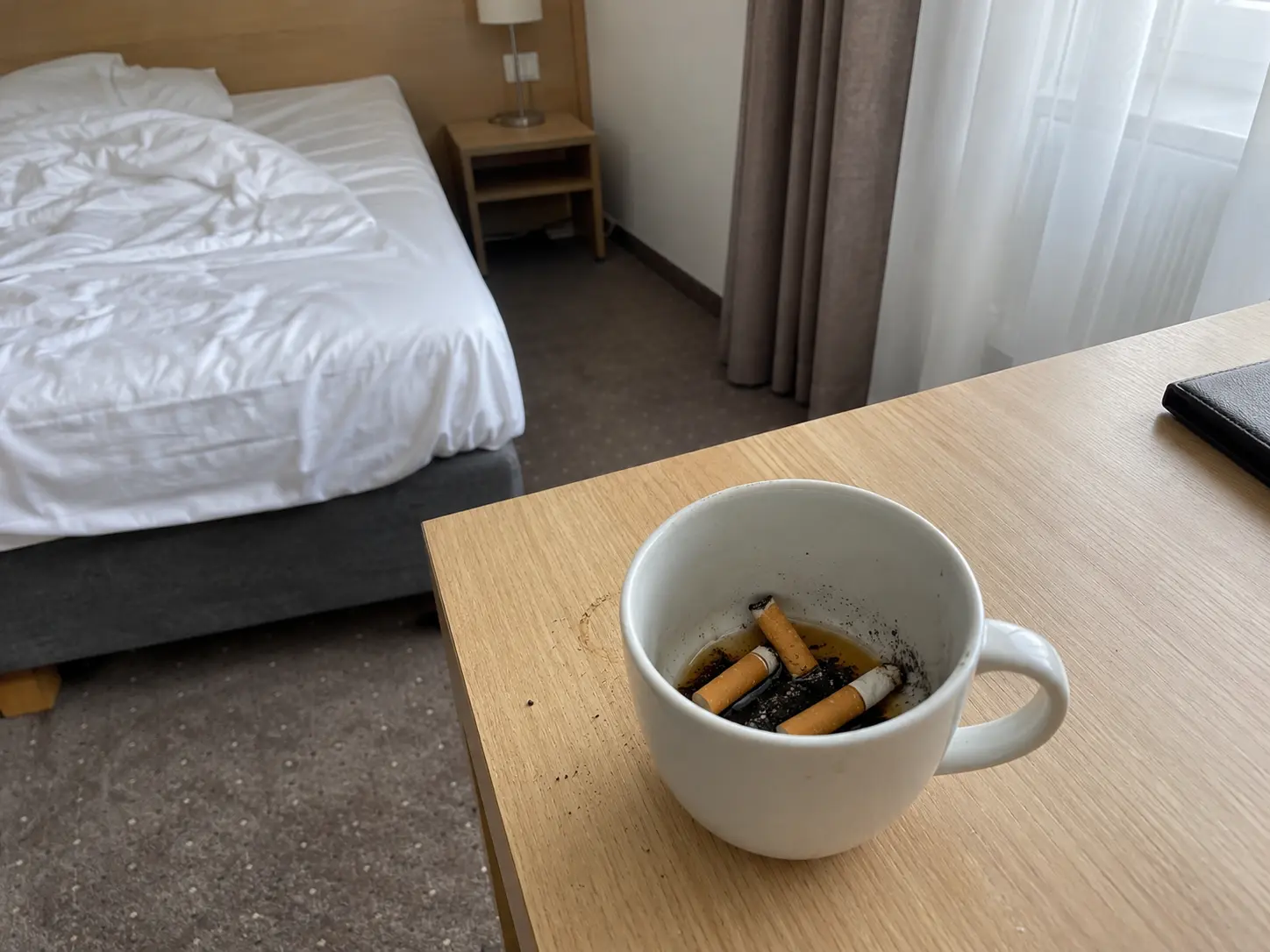 Ausgedrückte Zigarettenstummel und Asche in einer weißen Kaffeetasse auf dem Schreibtisch eines Hotelzimmers, dahinter das ungemachte Bett